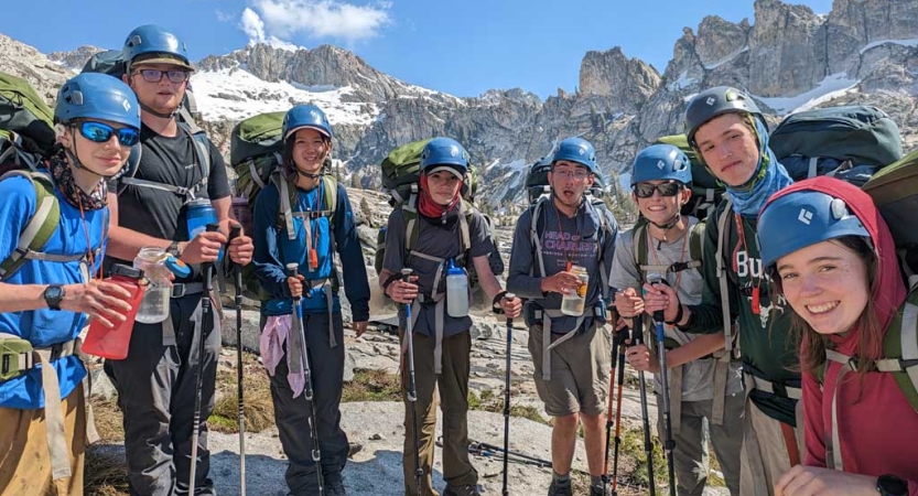 A group of students wearing helmets and backpacks stand in front of a mountainous ridge. 